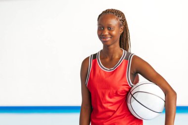 Yatay bir fotoğraf kırmızı üniformalı genç bir bayan basketbolcu kapalı bir sahada kolunun altında basketbol topunu tutarken özgüvenle gülümsüyor ve spora olan tutkusunu ve coşkusunu vurguluyor.