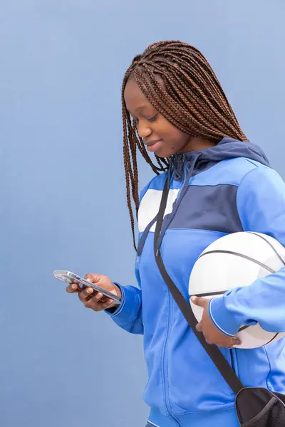 stock image Vertical photo a young woman in blue sportswear holds a basketball and looks at her smartphone while smiling. She carries a black sports bag over her shoulder and stands against a blue background.