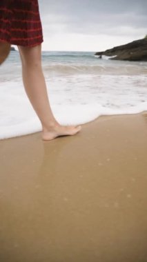 Yavaş çekim dikey video 1080, kumlu bir plajda çıplak ayakla yürüyen kırmızı elbiseli bir kadını yakalar. Okyanus dalgalarının rahatlatıcı sesi arka planı doldurur. Seyahat kavramı.