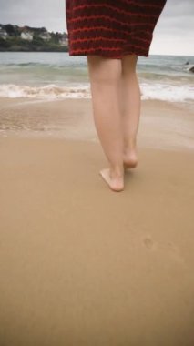 Kumlu bir sahilde çıplak ayakla yürüyen kırmızı elbiseli bir kadının yavaş çekim dikey videosu. Okyanus dalgalarının rahatlatıcı sesi rahatlatıcı bir arka plan sağlar. Seyahat kavramı.