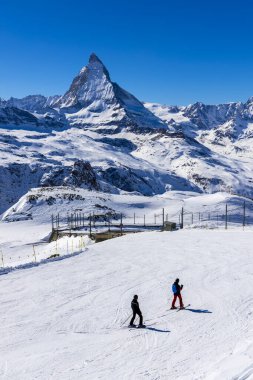 Two skiers on the way to ski slope in Swiss Alps in sunny day,Matterhorn behind. Small wooden house near by ski slope.Mountains and skiing resort for winter travel.Winter landscape of Switzerland.