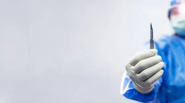 Mavi elbiseli bir erkek doktor ya da cerrah elinde cerrahi neşter ya da bulanık beyaz arka planda tıbbi bıçak tutuyor. Kozmetik ya da ortopedik ameliyat için cerrahi bıçak. Açık renk hoparlör takan adam..