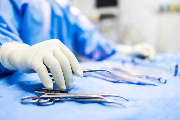 Hand of doctor or surgeon in blue gown pick up surgical clamp instrument or tools inside operating theatre with blur background.Medical equipment for surgery inside room in hospital.Modern technology.
