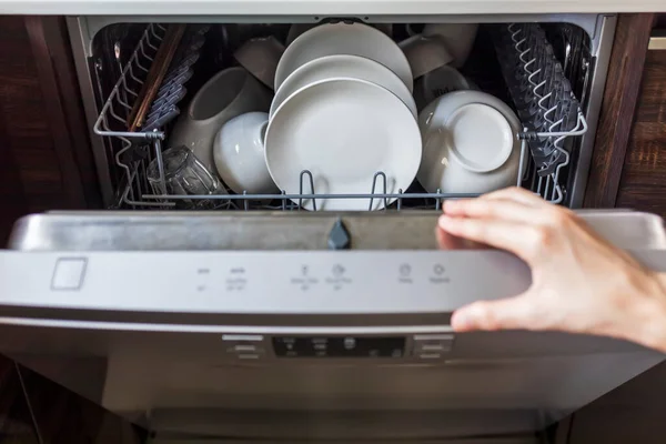 Kadın kullanılmış tabağı evde, mutfakta bulaşık makinesinde dolduruyor. Rafı tutan kadının eli. Çamaşır makinesinin içinde beyaz, temiz tabak ya da kap..