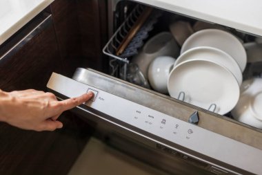 Kadın kullanılmış tabağı evdeki mutfakta bulaşık makinesinde dolduruyor. Kadının eli düğmeye basıyor. Çamaşır makinesinin içinde beyaz, temiz tabak ya da bardak var..