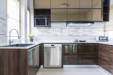 Ahşap ve cam dolaplı modern mutfak tasarımı. Su lavabosu ve musluğu beyaz masanın üstünde. Elektrikli bulaşık makinesi ve fırın. Mikrodalga ve fırın kaputu..