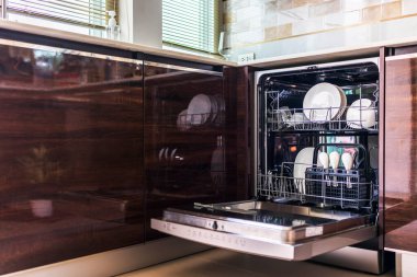 Bulaşık makinesi güzel mutfakta, yıkanmaya hazır beyaz kullanılmış bulaşıklarla. Ahşap mutfakta elektronik bulaşık makinesi, pencerelerden gelen ışık. Kapısı açık ev eşyaları teknolojisi..