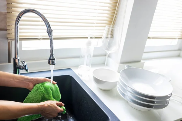 Birçok temiz beyaz tabak mutfakta beyaz mermer bir masanın üzerinde bulanık bir arka planı var. Ev hanımı siyah lavaboda muslukla temizlik bezi yıkıyor. Bulaşık deterjanı ile pencereyi yıkıyor..