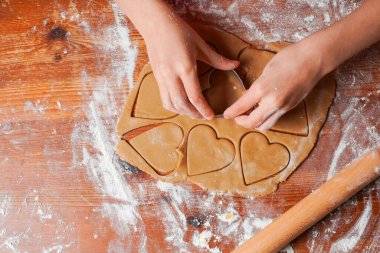 Hands of child makes biscuits cookies as hearts out of dough. Rolling pin and flour on a table. Valentines Day celebration.