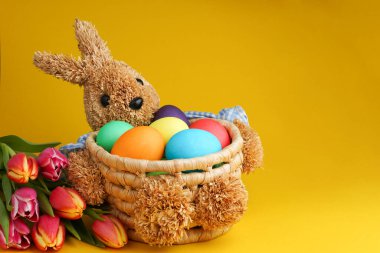 Straw bunny basket with easter colorful eggs und fresh tulips on yellow background. Happy Easter card. Close up.