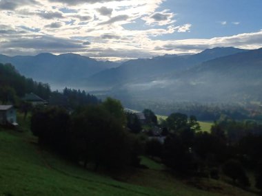 Haute-Savoie, Fransa 'daki Giffre Vadisi' nin nefes kesici manzarası. Verdant tarlaları, yumuşak bulutlar ve güneş ışığıyla dağların sisli katmanlarına çıkar ve bu huzurlu dağlık alana derinlik katar. Doğa, seyahat ve manzara temaları için mükemmel