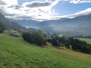 Haute-Savoie, Fransa 'daki Giffre Vadisi' nin nefes kesici manzarası. Verdant tarlaları, yumuşak bulutlar ve güneş ışığıyla dağların sisli katmanlarına çıkar ve bu huzurlu dağlık alana derinlik katar. Doğa, seyahat ve manzara temaları için mükemmel