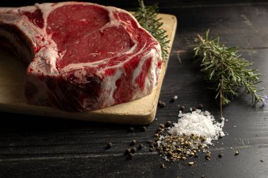 A raw beef chop on a chopping board with herbs and seasoning on a dark background