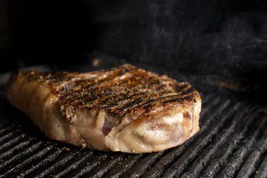 A beef chop cooking on a barbeque with smoke, on a dark background