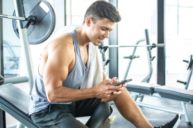 Handsome young man resting on weight bench and using phone after training in a gym.