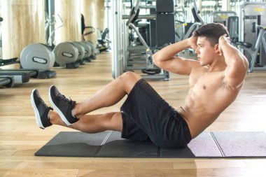 Handsome shirtless man doing crunches on exercise mat in gym. Young sporty guy doing crunches with hands on head in gym.