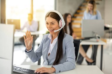 Smiling female operator agent with headset waving hand during video call on computer while having online consultation with customer at call center.
