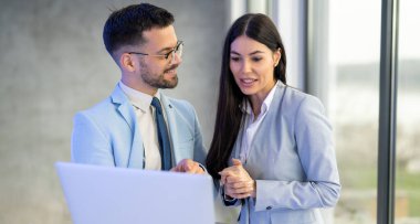 Yakışıklı bir iş adamı, ofis penceresinin yanında dururken bilgisayarında bayan meslektaşına bir şey gösteriyor.