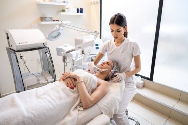 Güzellik uzmanı güzel genç kadın yüzüne spatulayla kozmetik jel soyma maskesi uyguluyor. Cilt bakımı ve kozmetoloji spa konsepti.
