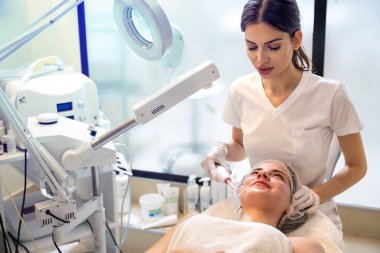 Kadın güzellik uzmanı güzellik kliniğindeki güzel bayana Darsonval yüz masajı yapmak için kozmetoloji elektriği de kullanıyor..