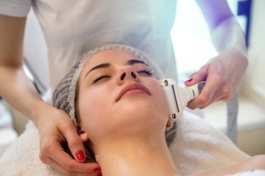 Güzellik salonunda elektrikli ultrason yüz masajı yaptıran genç bir kadın.