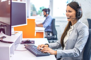 Gülümseyen yardımcı kadın operatör kulaklık ve mikrofon ile telefon görüşmesi sırasında müşteriyle konuşuyor ve bilgisayar veritabanındaki kesiciler hesabını kontrol ederek sorunu çözmeye çalışıyor.