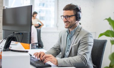 Orta yaşlı iş adamı modern bir ofiste kulaklık ve bilgisayar kullanıyor..