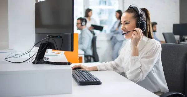 ヘッドセット付きの魅力的な若い女性従業員とコンピュータを使用して コールセンターで顧客をサポートするために話す — ストック写真