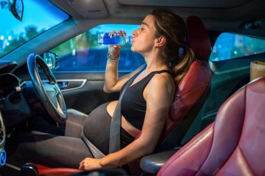 Arabada oturan ve plastik şişeden su içen hamile kadın trafik lambasının değişmesini bekliyor.