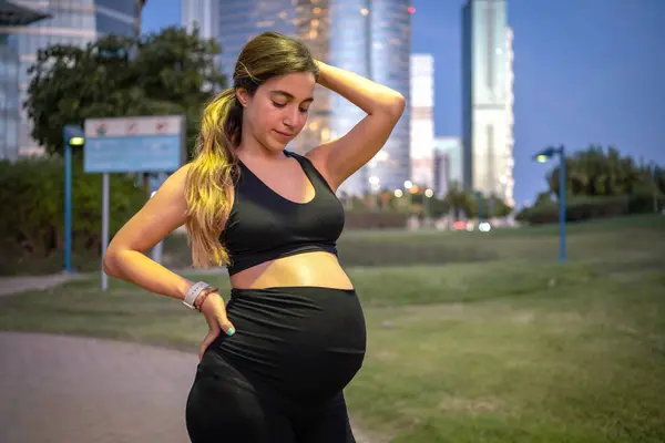 漂亮的年轻孕妇穿着黑色休闲装 晚上在户外摆姿势 看着自己9个月的肚子 — 图库照片