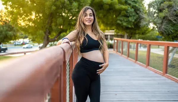 彼女の腹に触れ 屋外でカメラを見ながら 橋のフェンスの上に横たわる笑顔の妊婦 — ストック写真
