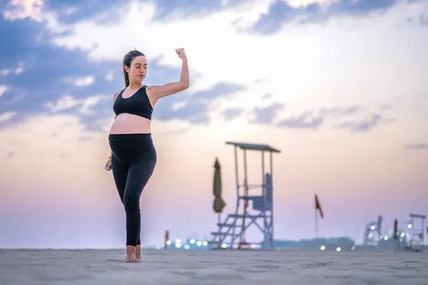 九个月的孕妇穿着运动服 在日落时站在沙滩上 表现出肌肉 健康生活方式 体育和怀孕概念 — 图库照片