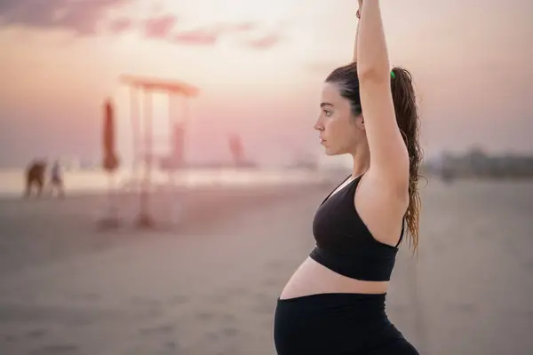 日落时 身穿黑色运动服的孕妇在海滩上摆姿势 使新月隆起瑜伽成为一个侧面景观 瑜伽和怀孕的概念 — 图库照片
