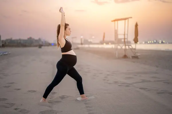 身穿黑色运动服的年轻孕妇在日落时在海滩上摆姿势 做新月隆起瑜伽 瑜伽和怀孕的概念 — 图库照片