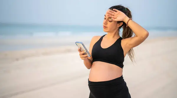 美しい妊娠中の女性が砂浜海岸の上に立って スマートフォンを見て 顔に疲れた握手を感じます — ストック写真