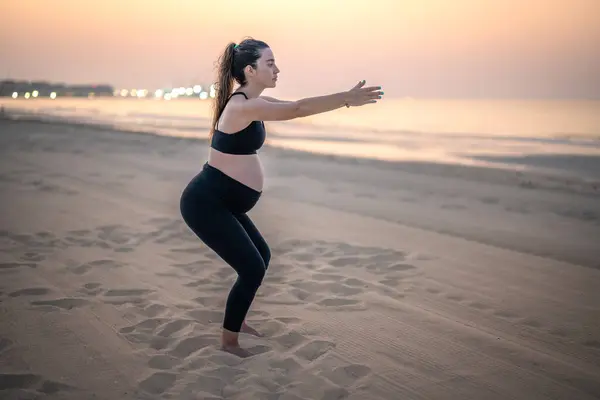 日落时一位年轻孕妇在沙滩上练蹲的全景 — 图库照片