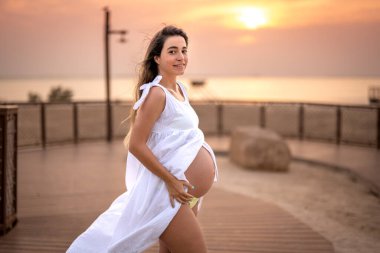 Dokuz aylık hamile bir kadın gün batımında beyaz elbisesiyle ahşap kaldırımda dikiliyor.