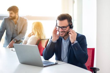 Kulaklıklı gülümseyen iş adamı laptopta çalışıyor, müşteriyle konuşuyor, çağrı merkezindeki çevrimiçi müşteri destek hizmetlerinde soruları cevaplıyor..
