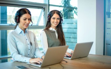 Kulaklık ve dizüstü bilgisayarlı iki kadın servis elemanı müşterilere yardım etmeye hazır.