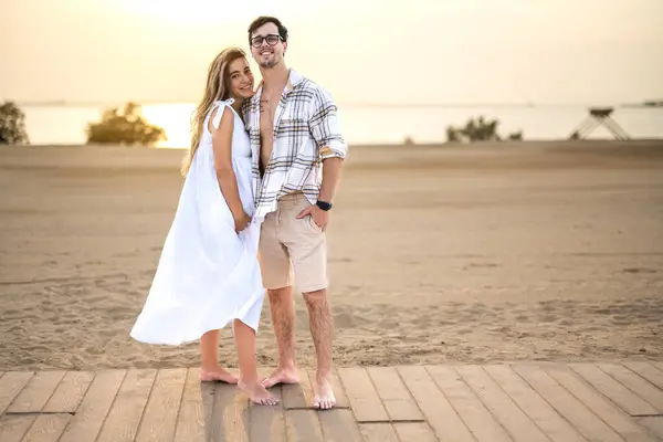 海岸に立っているカジュアルなウェアのカップルは 愛と抱き合い カメラを見ています 妊娠中の女性と歩道で抱擁するハンサムな男 — ストック写真
