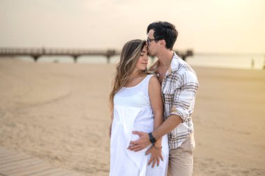 Kumsalda kucaklaşan mutlu bir çift. Yakışıklı bir adam hamile karısına sarılıyor ve gün batımında huzurlu anların tadını çıkarıyor.