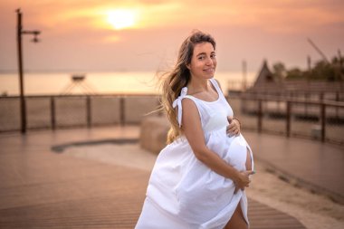 Dokuz aylık hamile kadın, beyaz elbiseler giyiyor ve gün batımında sahilde karnına dokunuyor.