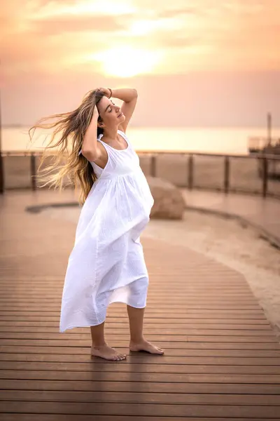 孕妇站在木制人行道上 享受着新鲜的海洋空气 日落时风吹拂着她的头发的全长肖像 — 图库照片