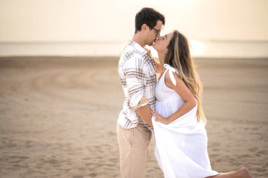 Sahilde öpüşen genç ve güzel çiftin yan görüntüsü. Beyaz elbiseli hamile bir kadın gün batımında kocasıyla kucaklaşıyor..