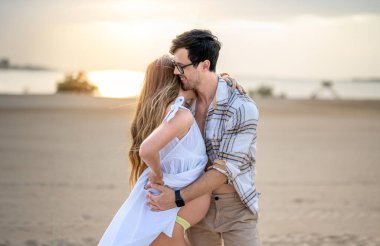 Yakışıklı genç adam günbatımında plajda hamile karısına sarılıyor.