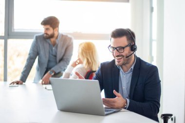 Kulaklık takmış gülümseyen işadamı laptopla çalışıyor, müşteriyle konuşuyor, ofisteki soruları cevaplıyor.