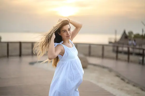 日没の海岸近くのビーチ遊歩道で風を楽しみながら白いドレスで美しい若い妊婦の肖像画 — ストック写真