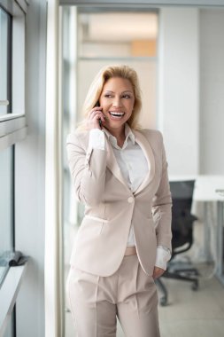 Parlak ofisin penceresinden bakarken telefonla konuşan olgun sarışın iş kadını.