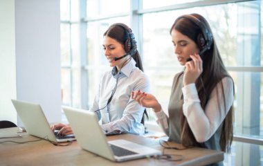 Aydınlık ofisteki telefon görüşmelerinde müşterilere yardımcı olan kulaklıklı ve dizüstü bilgisayarlı iki kadın servis operatörü..