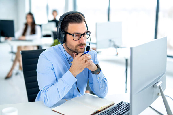 Pensive male customer service representative wearing eyeglasses and headset, holding pen with hand on chin and looking at computer screen.